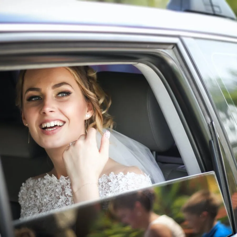 A bride is seen smiling in the back seat of a car.