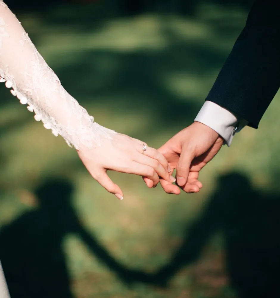 A close-up, blurred shot of a bride and groom holding hands. The bride, on the left, is wearing a white dress with lace sleeves and has a ring on her finger. The groom, on the right, is in a black suit. Their intertwined hands are in focus, with their shadows cast on the grass behind them.